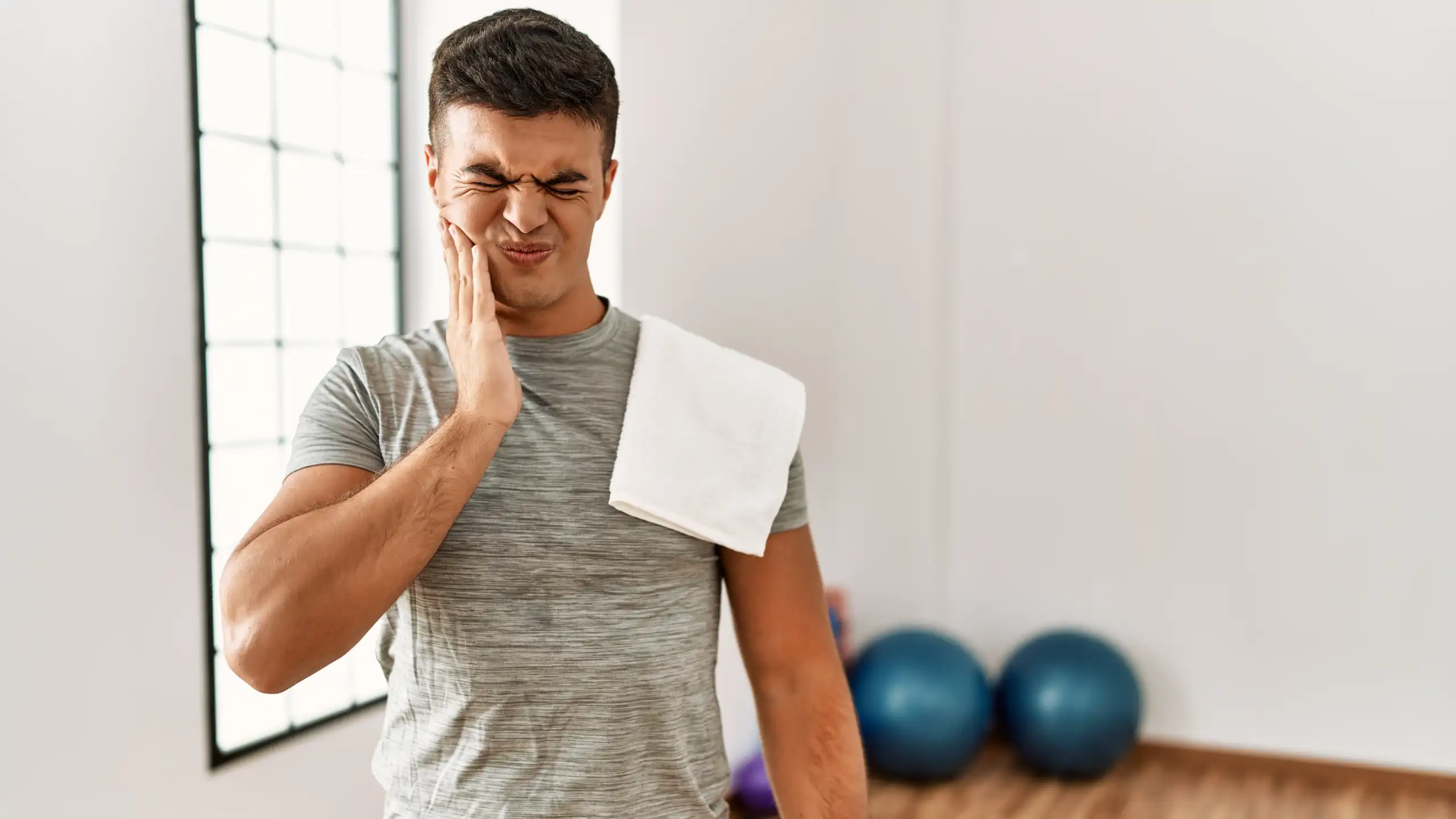 Ein junger sportlicher Mann in Trainingskleidung steht in einem Fitnessraum, hält sich schmerzverzerrt die Wange und zeigt Anzeichen von Zahnschmerzen. Ein Handtuch liegt über seiner Schulter, im Hintergrund sind Gymnastikbälle zu sehen.
