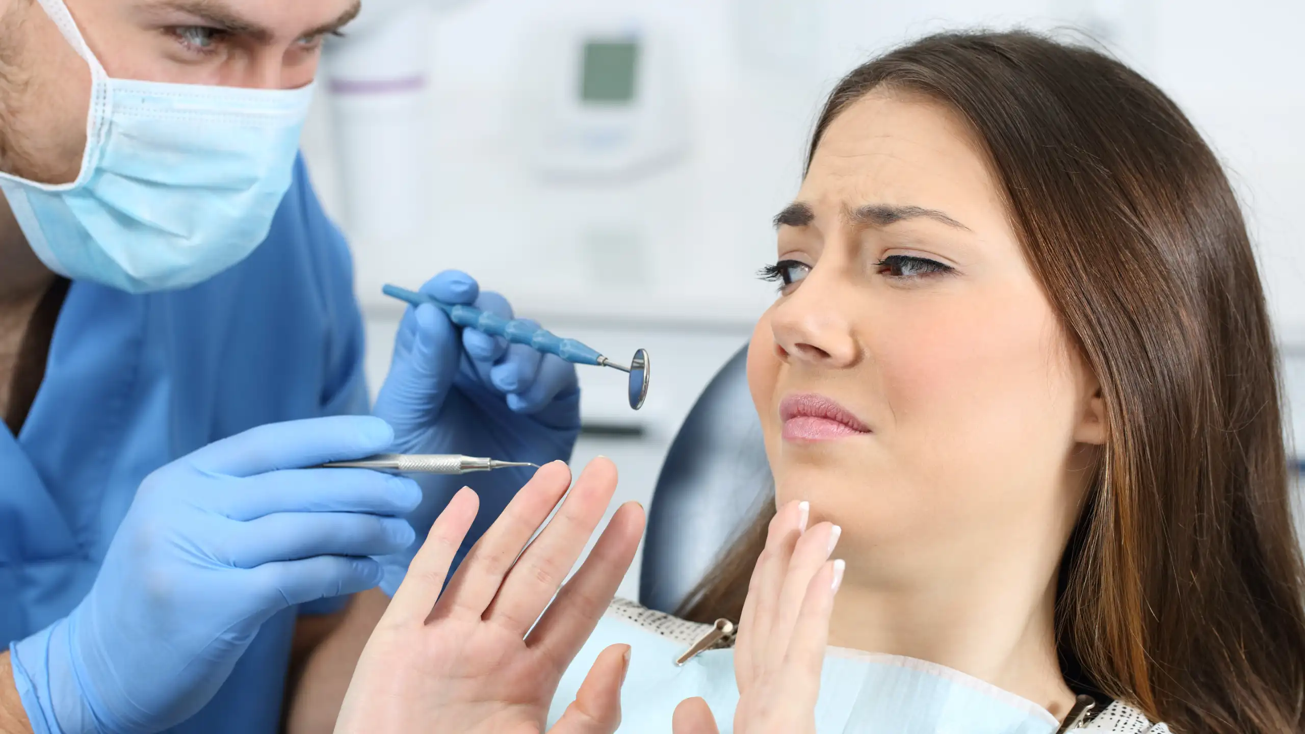 A woman in a dental chair looks anxious as a dentist with a mask and gloves holds dental tools close to her mouth.