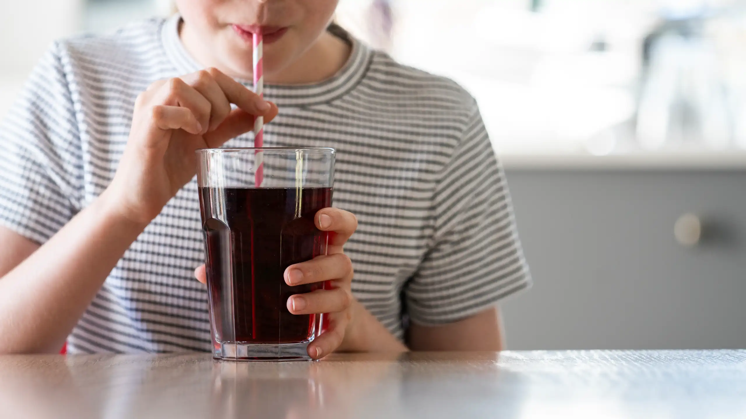 Person drinking a dark beverage through a striped straw from a glass, wearing a striped shirt, seated at a table.