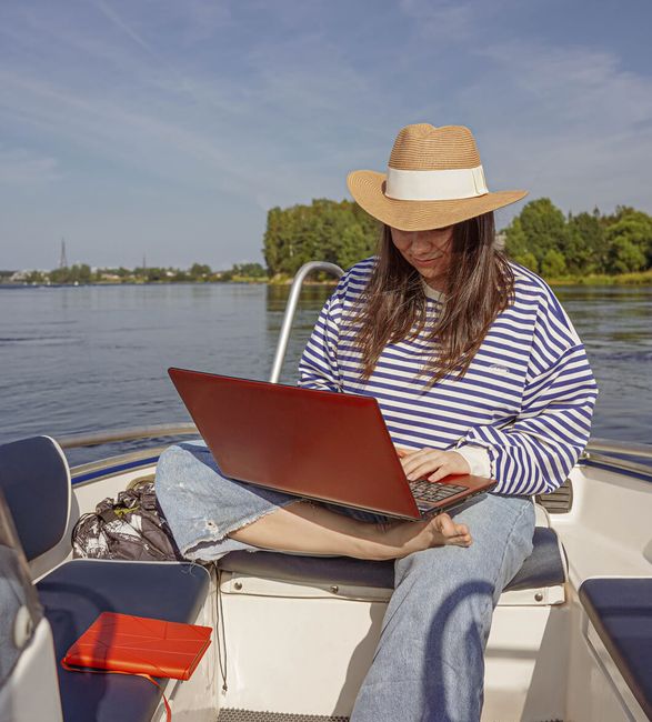 Woman on a boat using a laptop, with a serene water background.