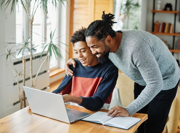 A smiling man leans over a teen, both looking at a laptop screen in a cozy room with plants and books.