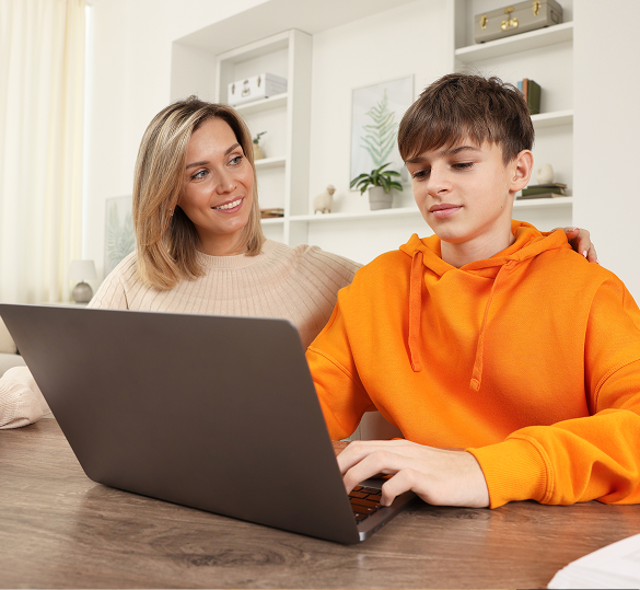 A woman smiles and supports a boy using a laptop at a table in a bright room with shelves and plants in the background.