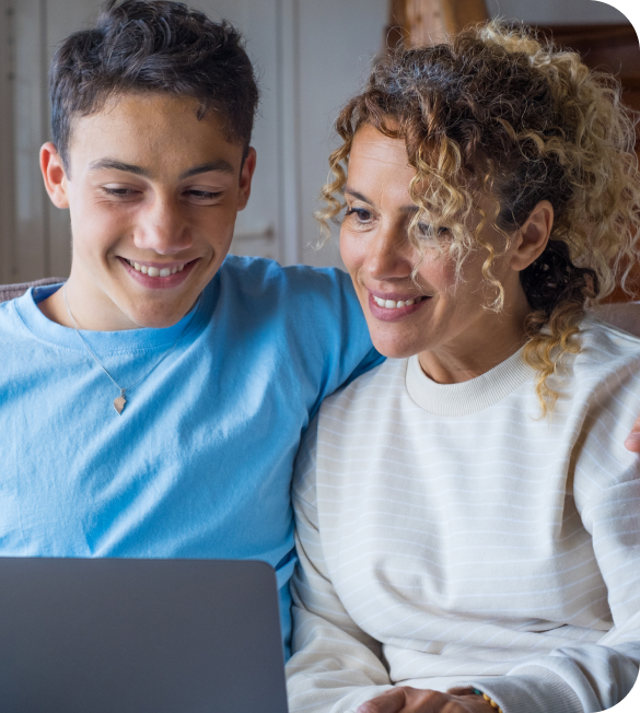 A smiling woman and a young man sit closely, looking at a laptop screen together in a cozy indoor setting.