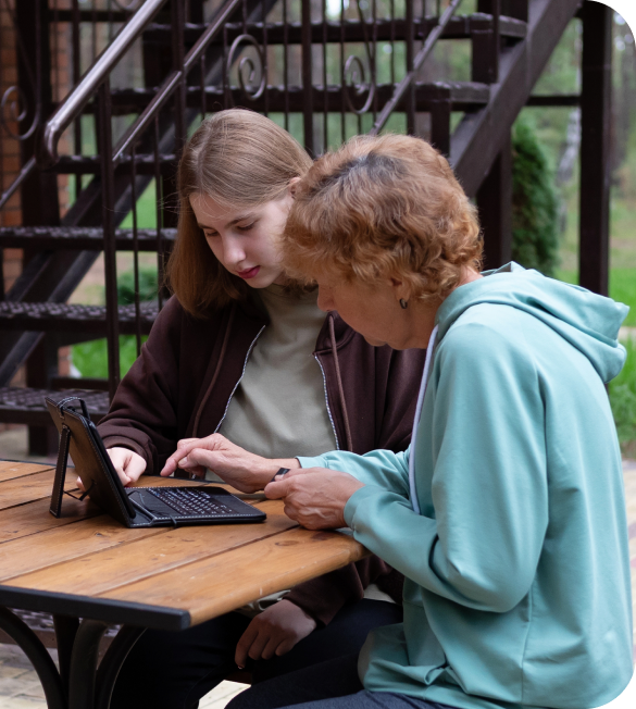 Two people seated at an outdoor table, focused on a tablet with a keyboard, surrounded by greenery and stairs in the background.