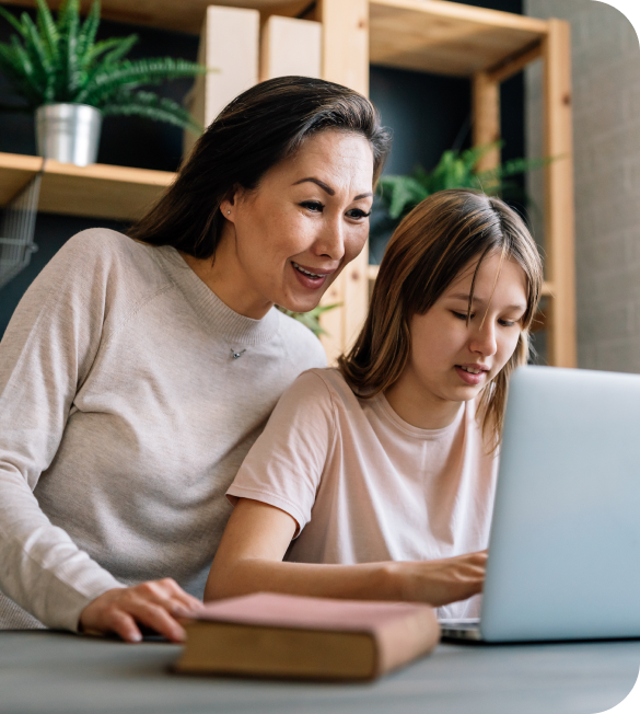 A woman and a girl are engaged with a laptop at a table, surrounded by shelves with plants and books in the background.