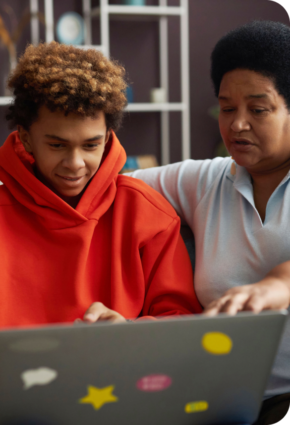A person in a red hoodie and another in a light blue shirt are sitting together, focused on a laptop with colorful stickers.