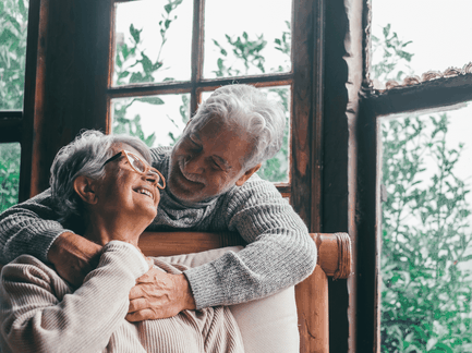 Happy elderly couple hugging and smiling in front of a large window overlooking greenery.