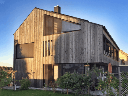 Modern detached house with wooden façade and large windows bathed in evening sunlight.