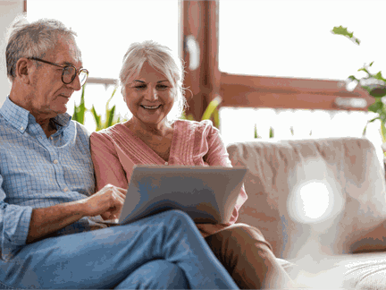 An elderly couple sits relaxed together on the sofa. Both are looking at a laptop.