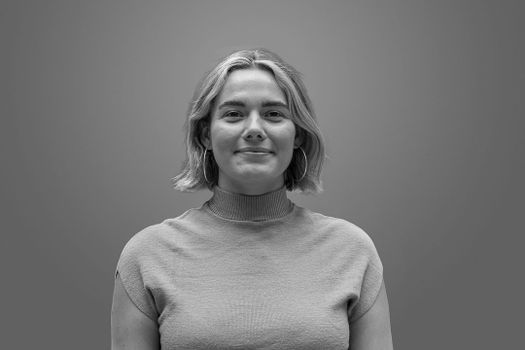 Asja Föllmi, in a black and white portrait of a person with short hair, wearing a turtleneck and hoop earrings, smiling slightly against a plain background.