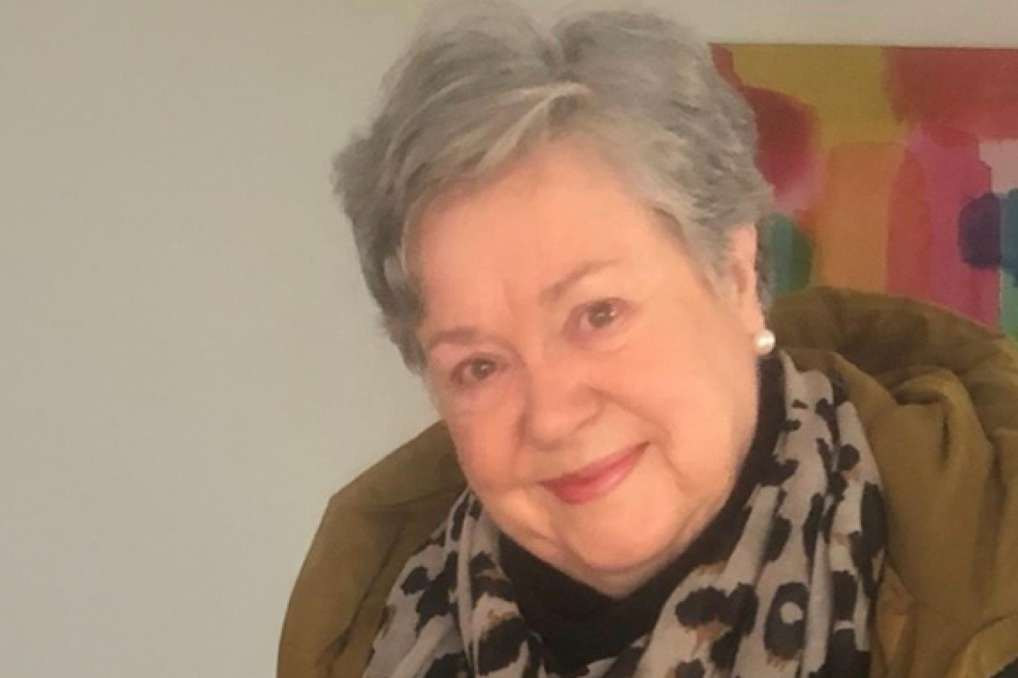 Older woman smiling, wearing a patterned scarf and a green jacket, with a colorful abstract painting in the background.