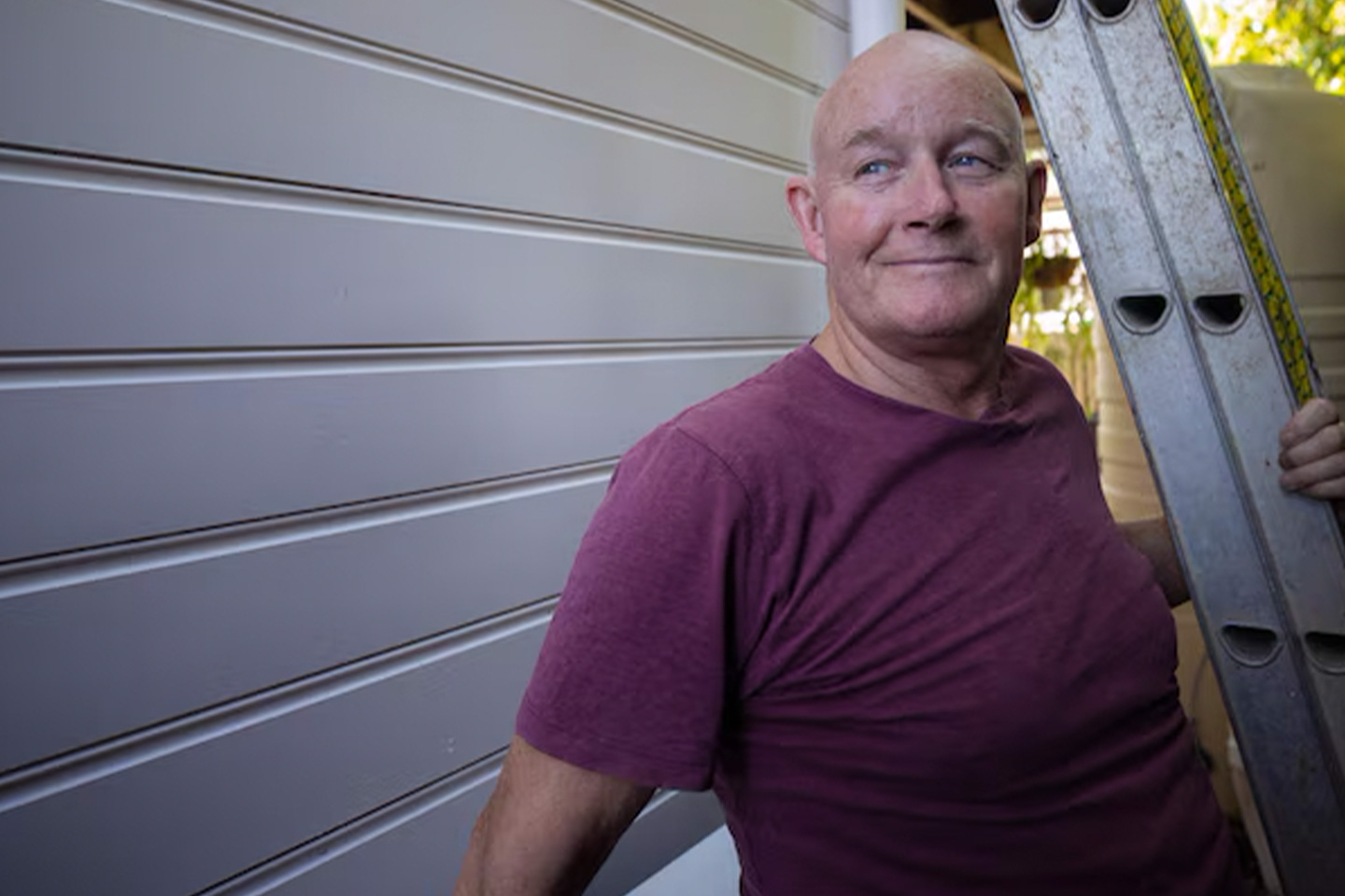 Photo of a smiling man in a maroon t shirt standing under a ladder