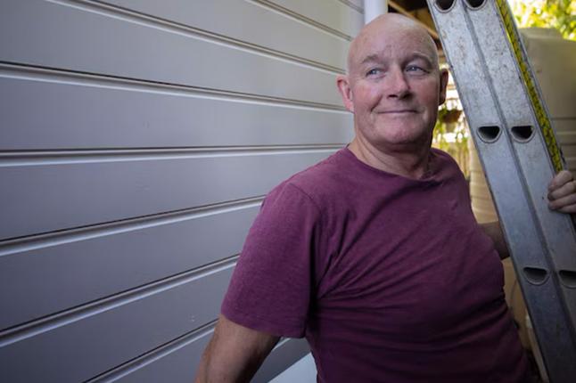 Photo of a smiling man in a maroon t shirt standing under a ladder