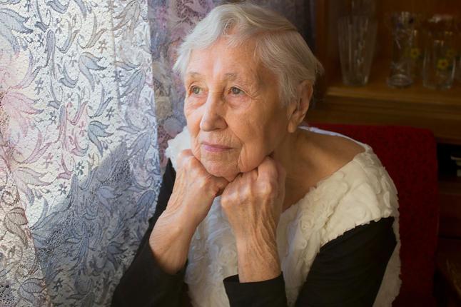 Image of an older woman with grey short hair looking out of a window in which the curtains are drawn. She also has both hands resting under her chin.