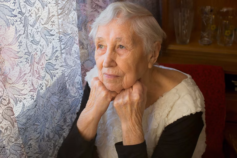 Image of an older woman with grey short hair looking out of a window in which the curtains are drawn. She also has both hands resting under her chin.