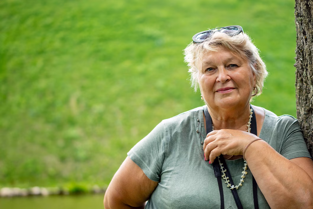 Image of an older Caucasian woman standing outside and leaning against a tree