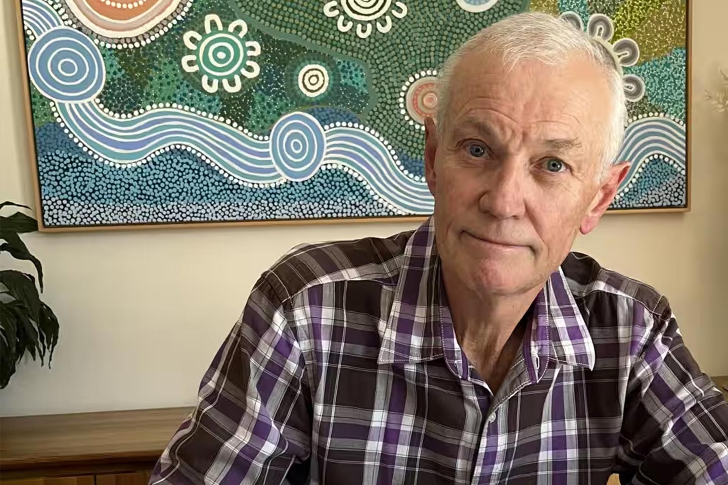 Photo of a man in a purple plaid shirt in front of an indigenous painting