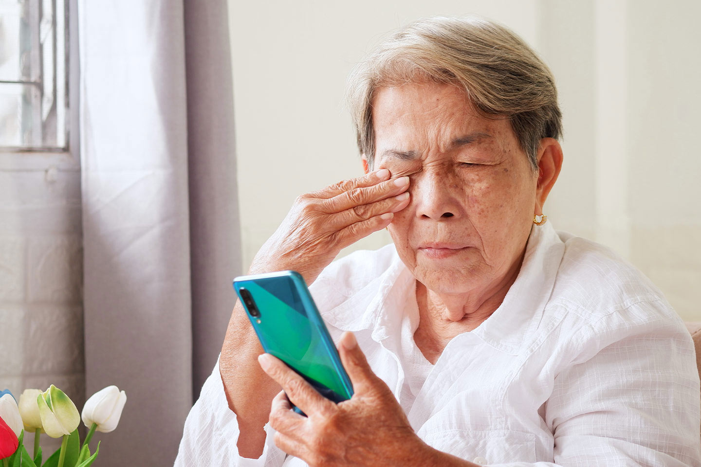 Older asian woman rubbing her eyes while looking at an smartphone