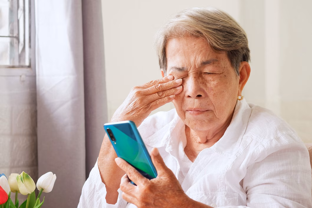Older asian woman rubbing her eyes while looking at an smartphone