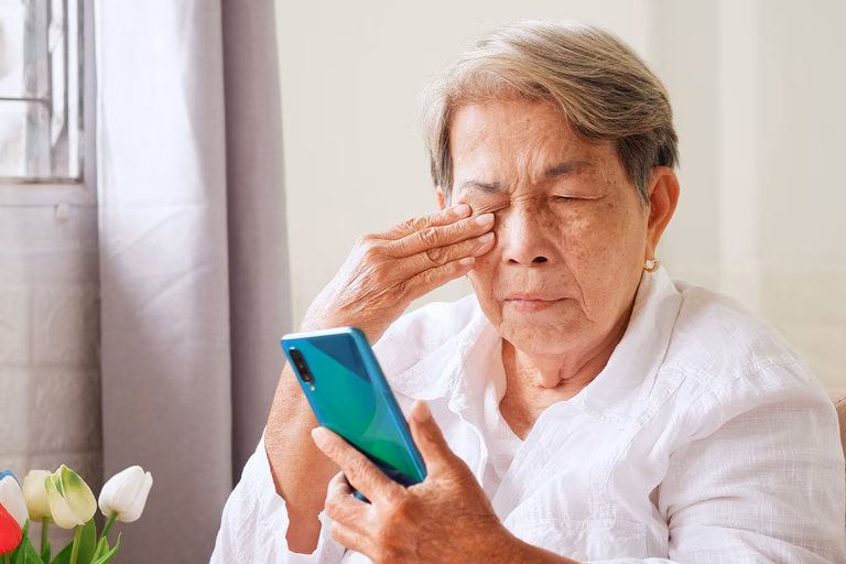Ältere asiatische Frau reibt sich die Augen, während sie auf ein Smartphone schaut