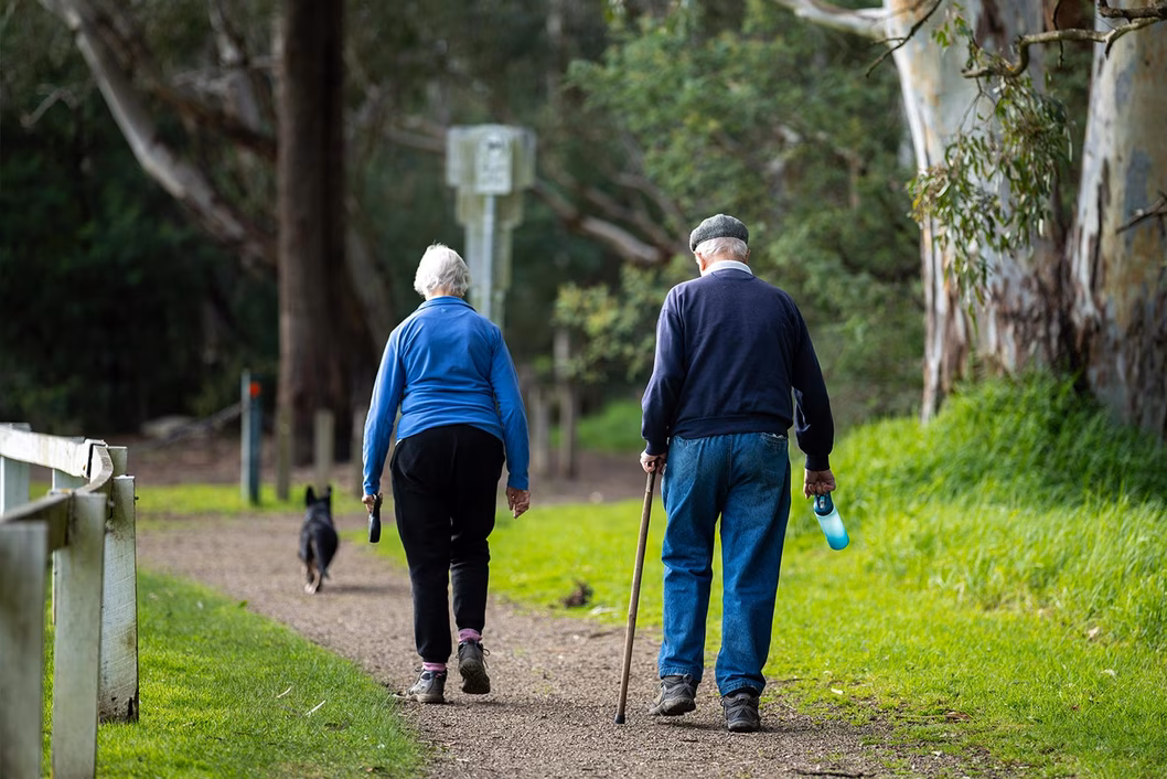 Image of an older man with a walking stick and an older woman walking with their dog along a park track