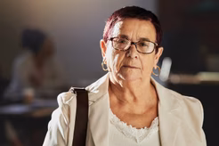 Image of an older woman with short dark hair, glasses and a hand bag strap over her right shoulder