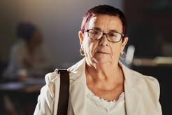 Image of an older woman with short dark hair, glasses and a hand bag strap over her right shoulder