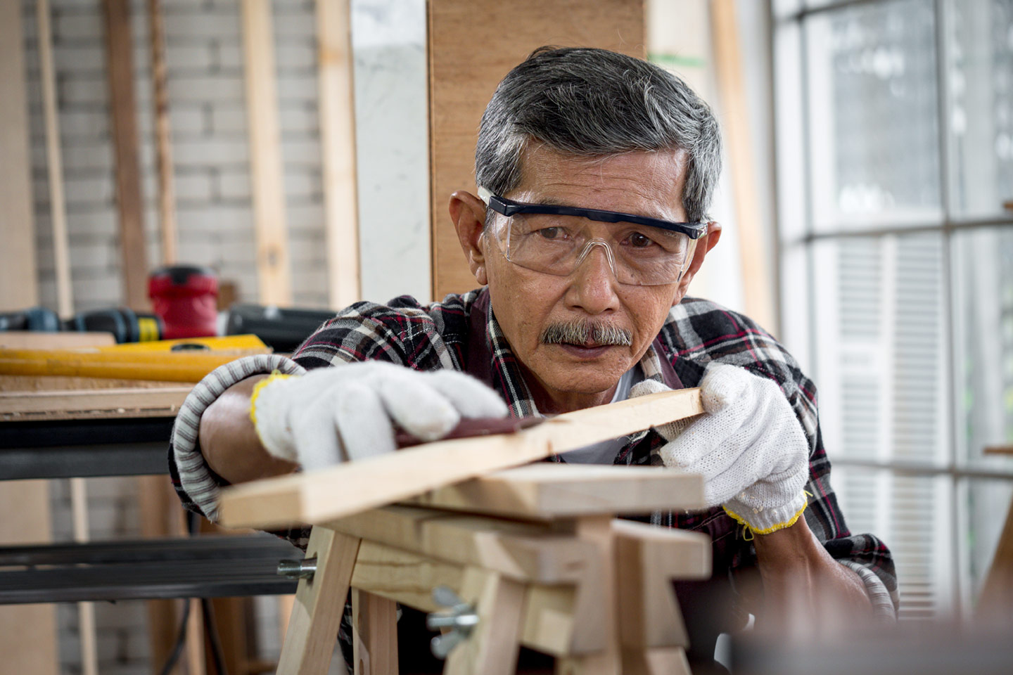 Older Asian carpenter wearing goggles working on a piece of timber