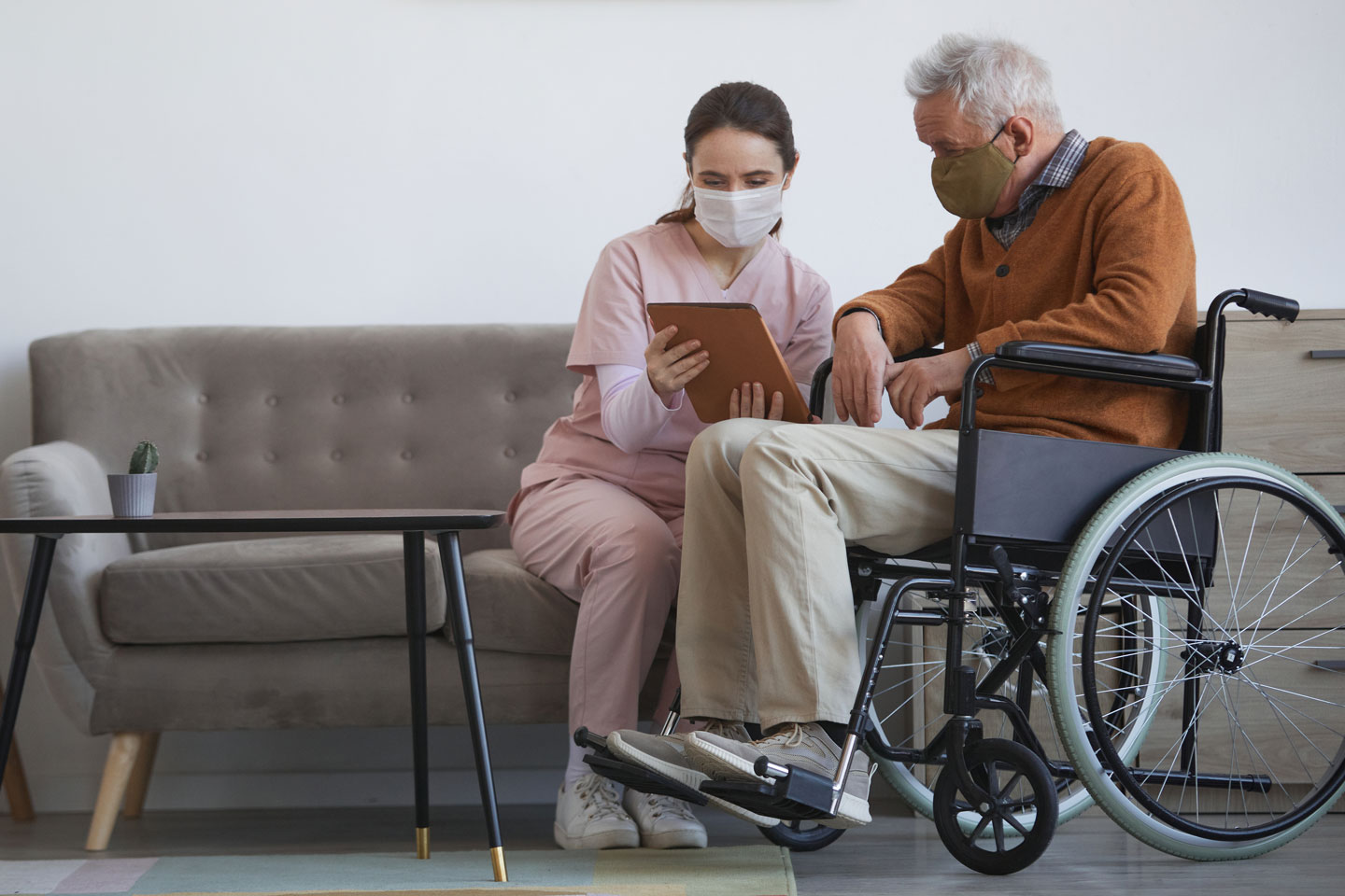 Picture of older man in wheelchair wearing mask listening to nurse on couch wearing mask
