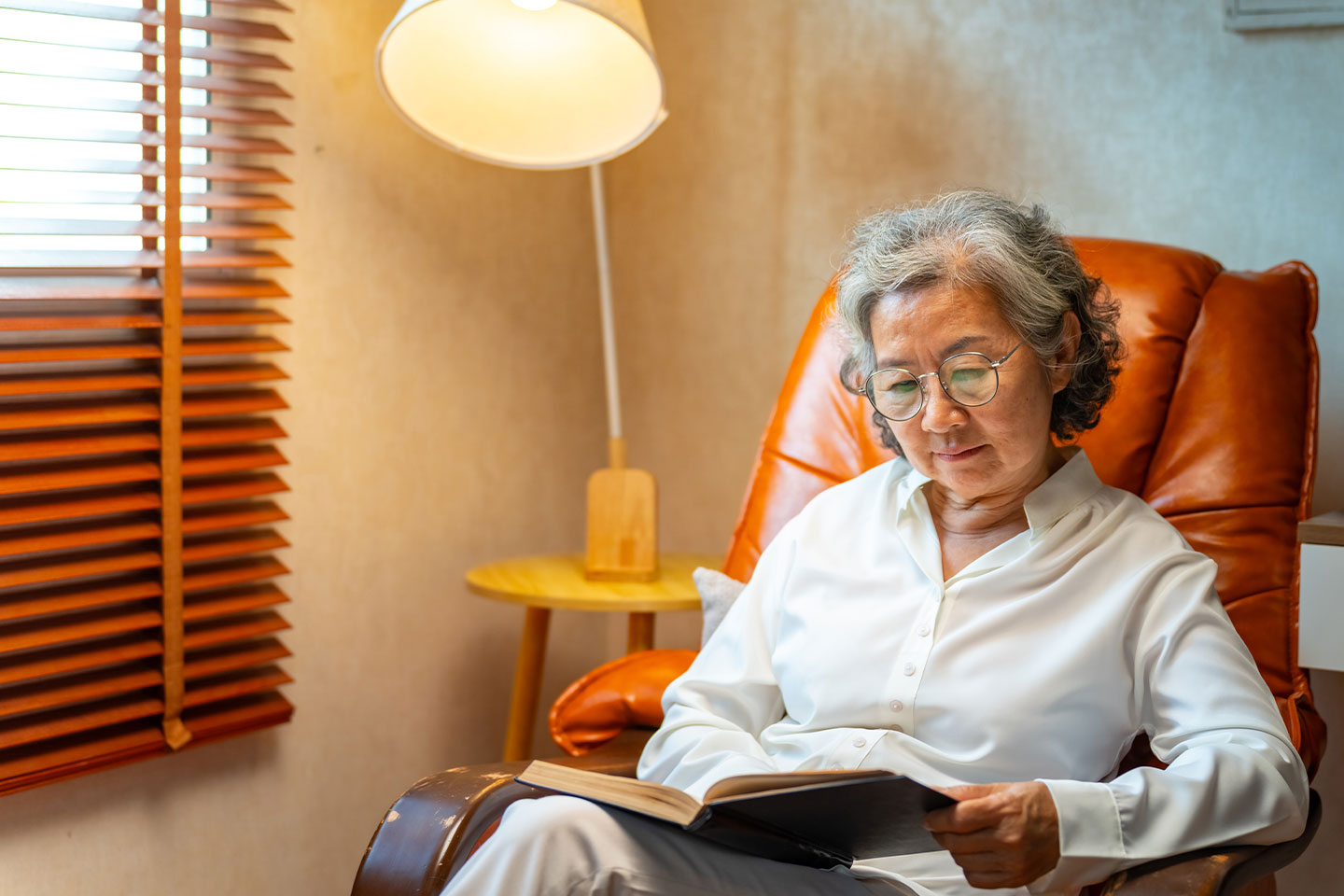 Image of an older Asian woman sitting in an armchair reading a book