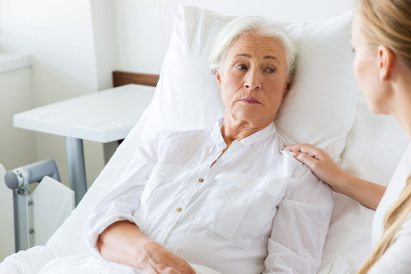 Photo of a woman in white in a hospital bed with a nurse attending