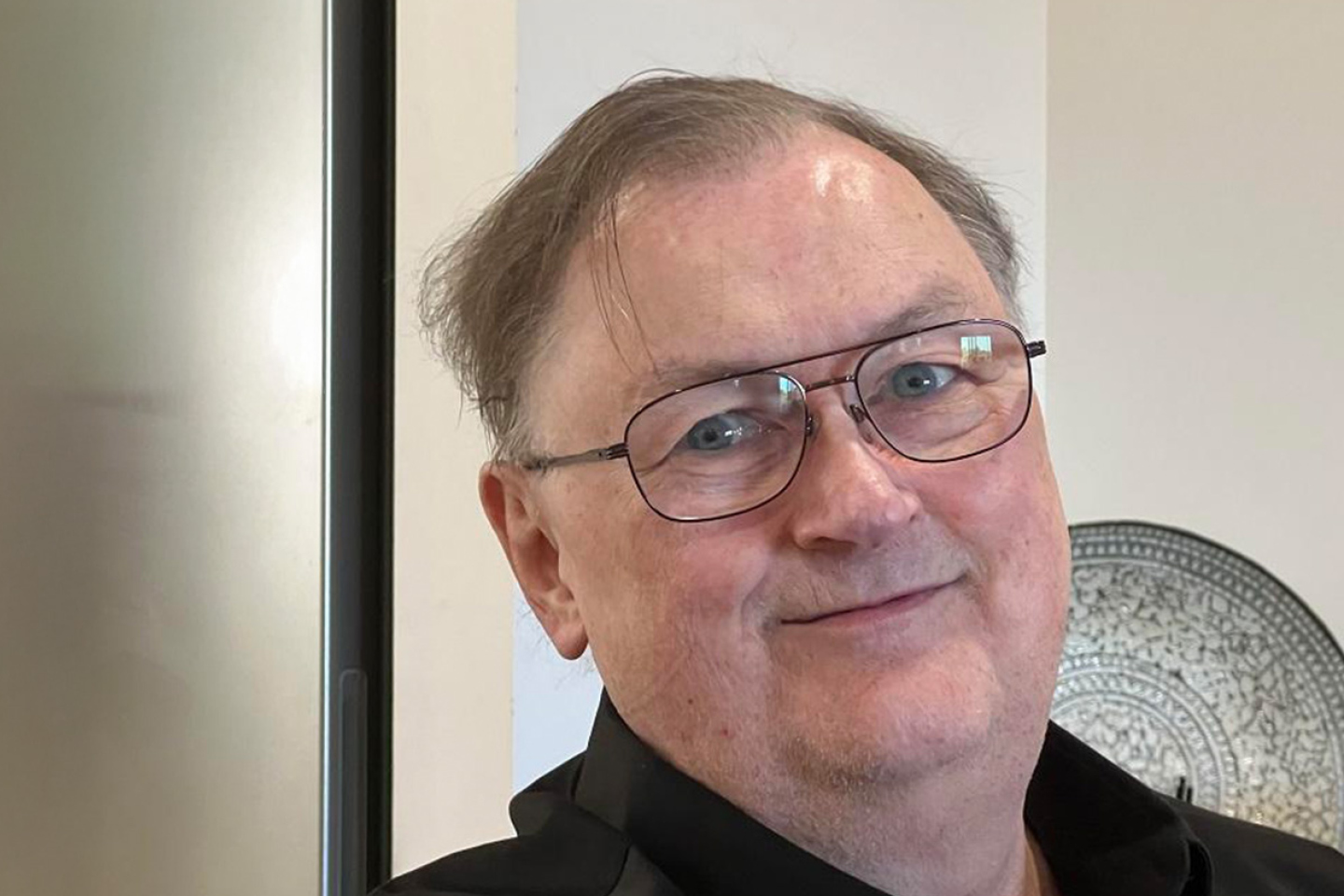 Smiling person with glasses and short hair, wearing a black jacket, stands indoors near a metallic decorative plate.