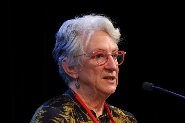 An elderly person with gray hair, red glasses, and a patterned outfit speaks at a microphone against a dark background.
