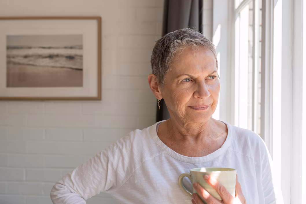 Im age on an older woman in a white long sleeve t-shirt hold a mug, looking out of a window