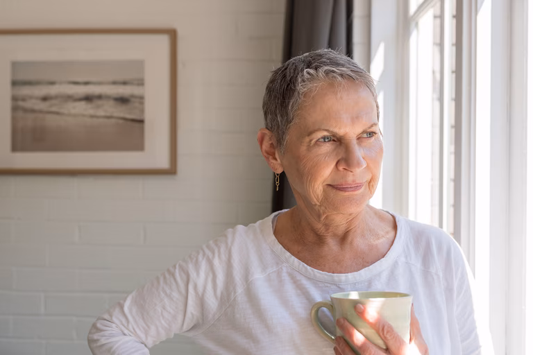 Im age on an older woman in a white long sleeve t-shirt hold a mug, looking out of a window