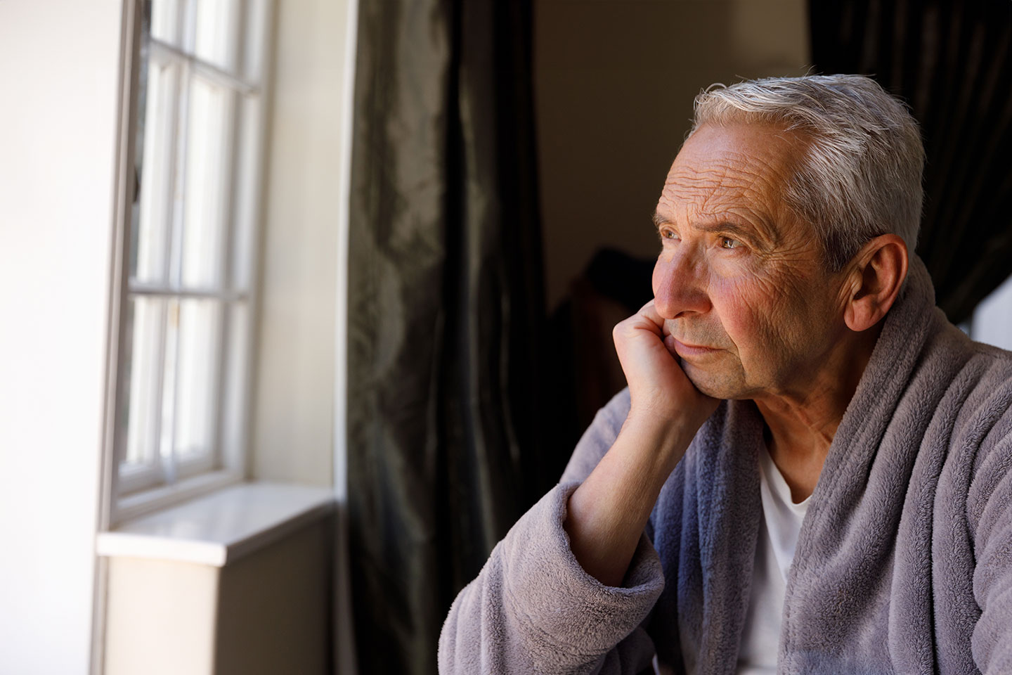 Image of an older man wearing a dressing gown and looking out of a window