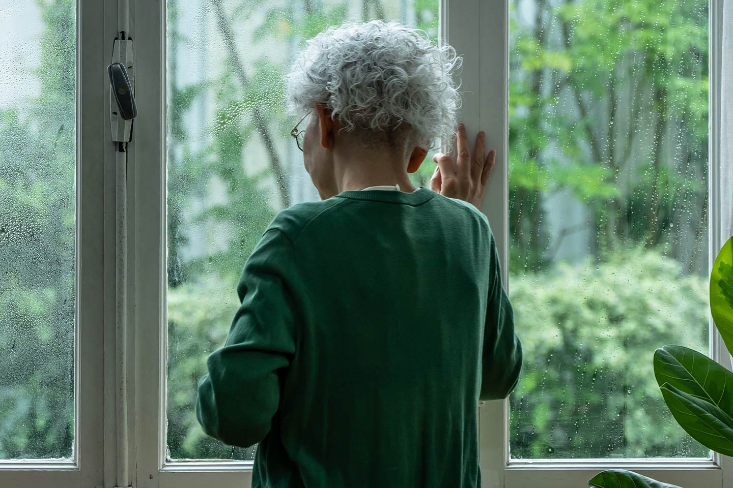 Image of an older asian woman looking downcast out a window. She is wearing a racing green pullover and it is raining outside.