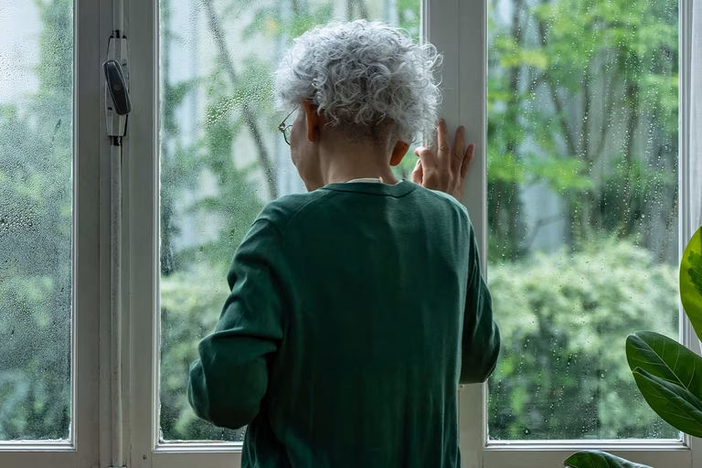Bild einer älteren asiatischen Frau, die niedergeschlagen aus dem Fenster schaut. Sie trägt einen rennsportgrünen Pullover und draußen regnet es.