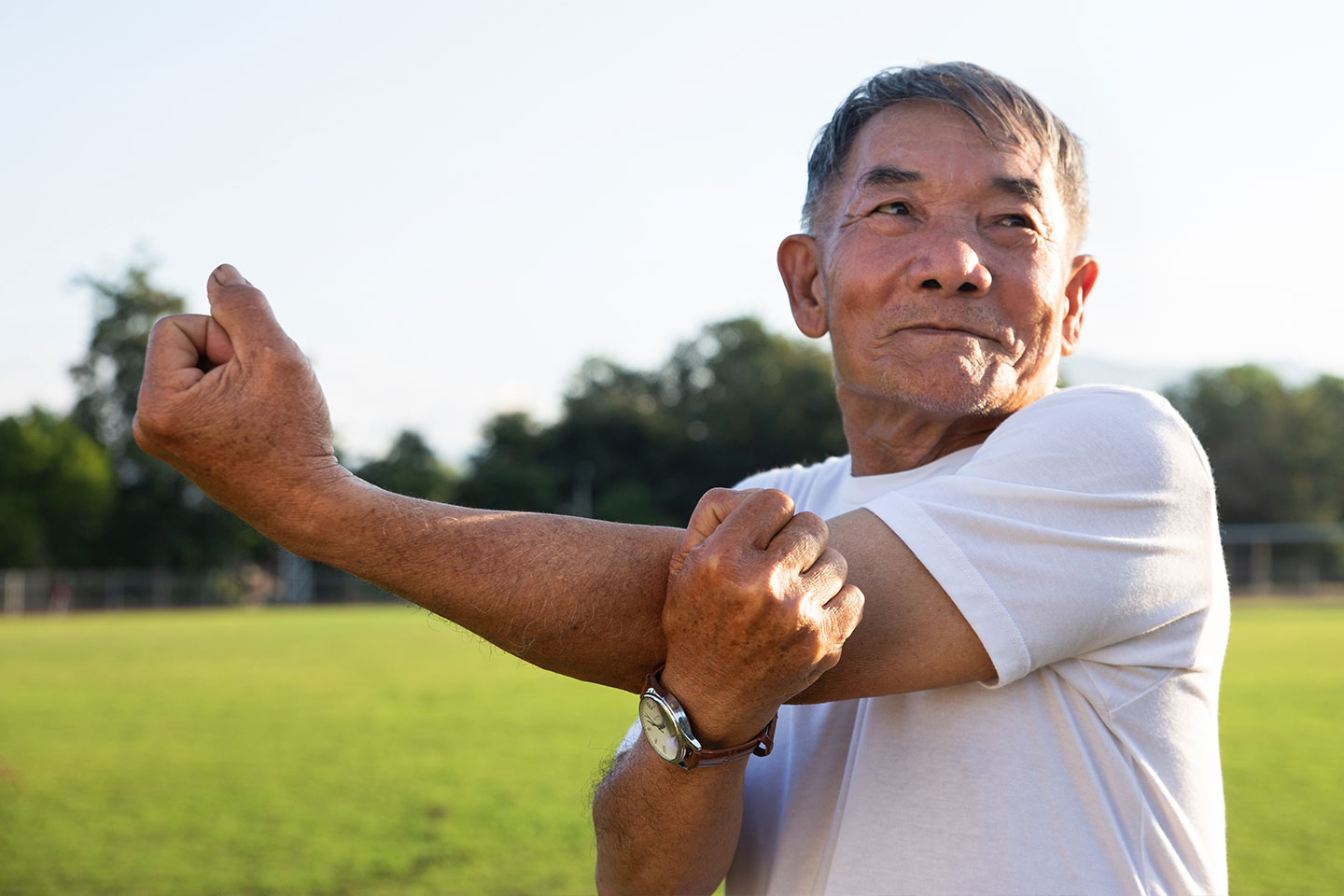 Asian older man stretching outside in a park oval on a sunny day