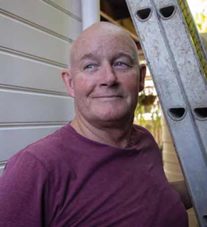Photo of a smiling man in a maroon t shirt standing under a ladder