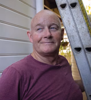 Photo of a smiling man in a maroon t shirt standing under a ladder