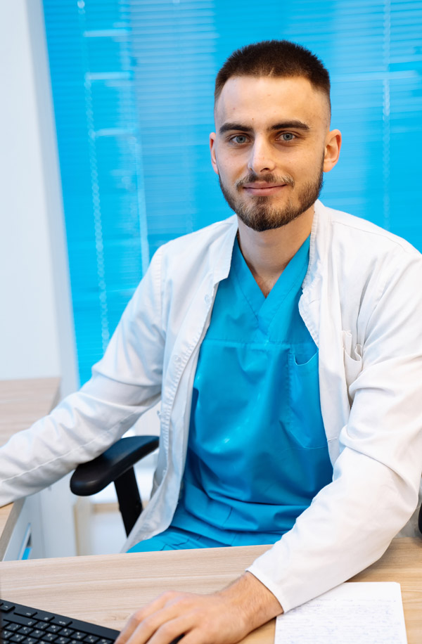 Young man in medical uniform at computer looking to camera