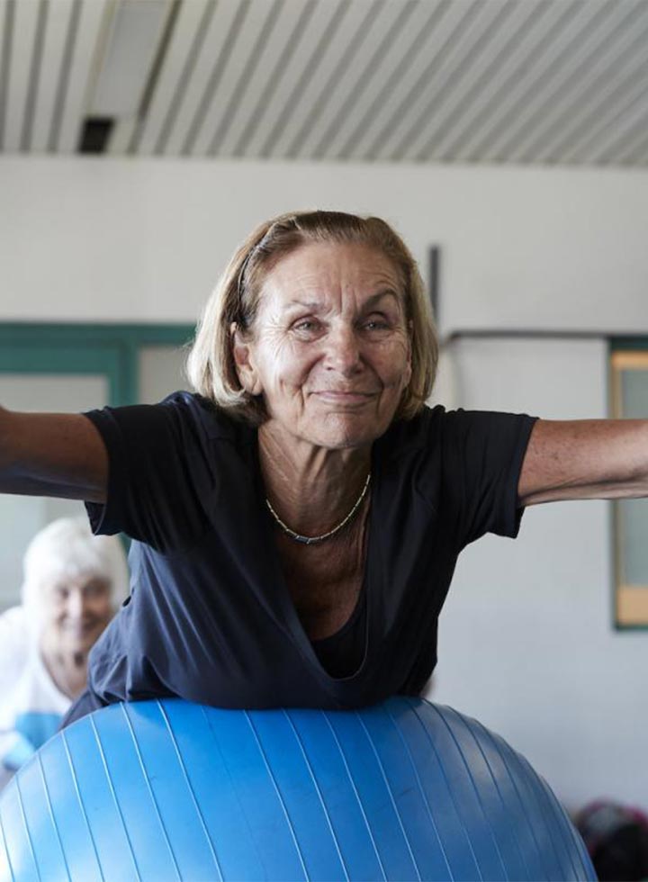 Image of an older woman exercising indoors with a group of older woman