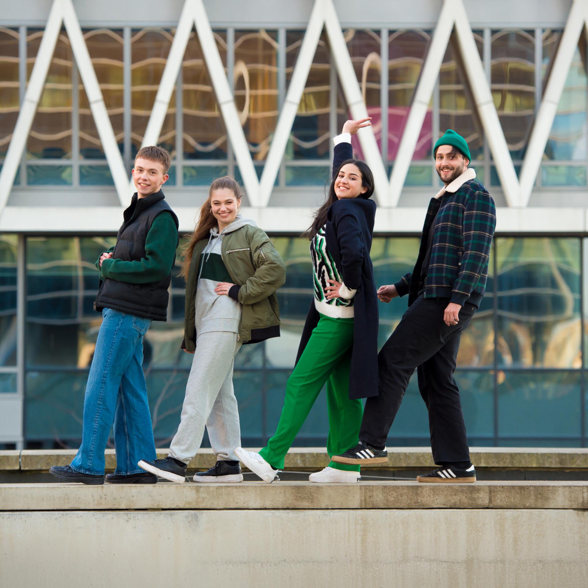 Four people posing playfully in front of a modern building with geometric design, dressed casually in colorful attire.