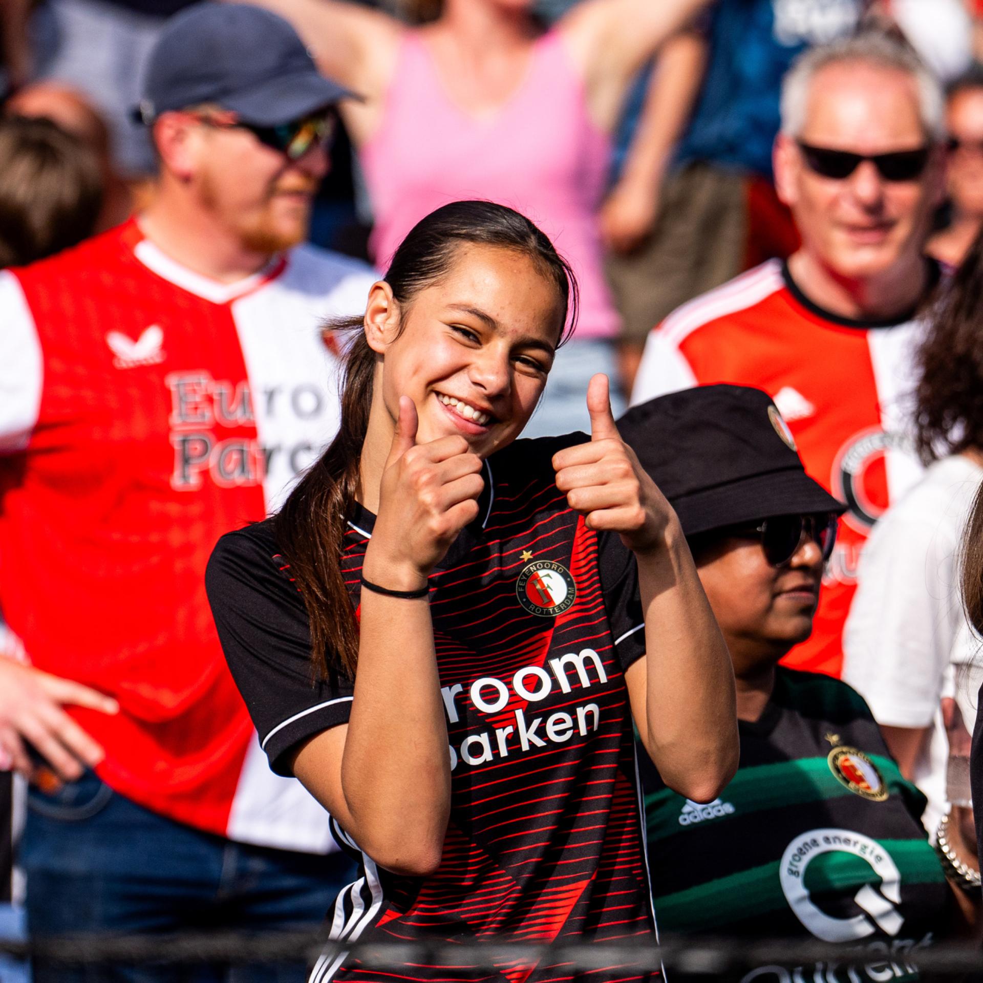 Young woman smiling and giving thumbs up in a crowd, wearing a red and black sports jersey, surrounded by other fans in similar attire.