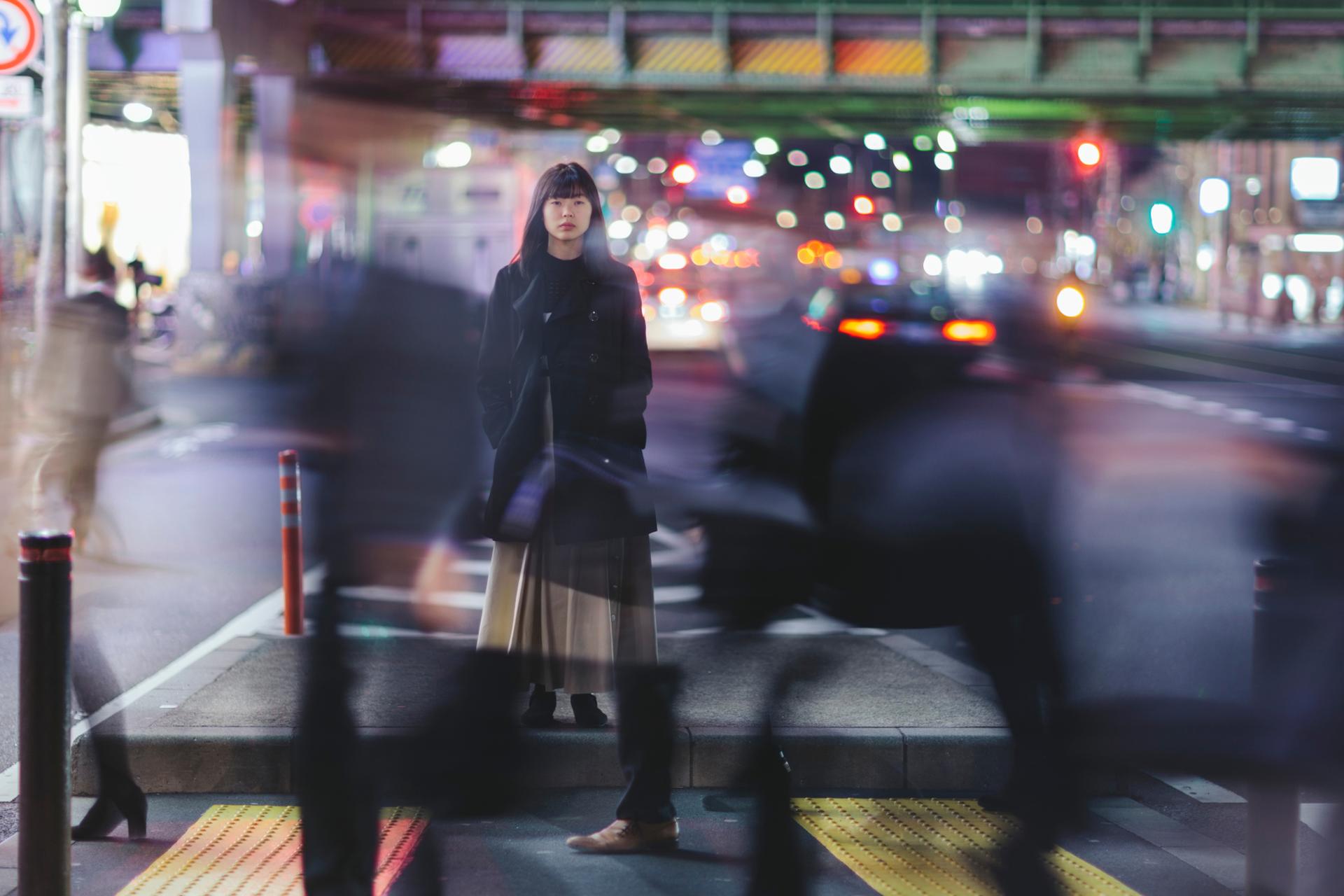 A woman stands still on a city street at night, surrounded by blurred motion of people and cars, with colorful lights in the background.