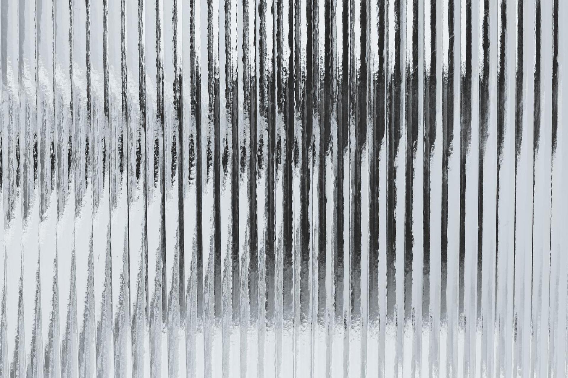 A close-up of a textured, corrugated glass surface with vertical ridges, creating a distorted reflection of light and shadow.