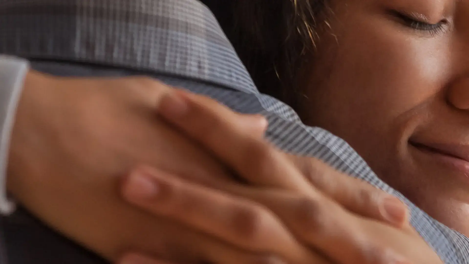Close-up of two people hugging, showing one person's hand on the other's shoulder. Both have content expressions.
