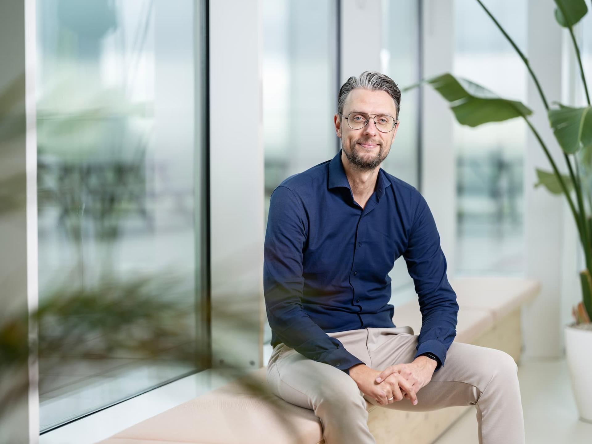 Danny van Steijn in a navy shirt and beige pants sitting on a bench indoors, with large windows and plants in the background.