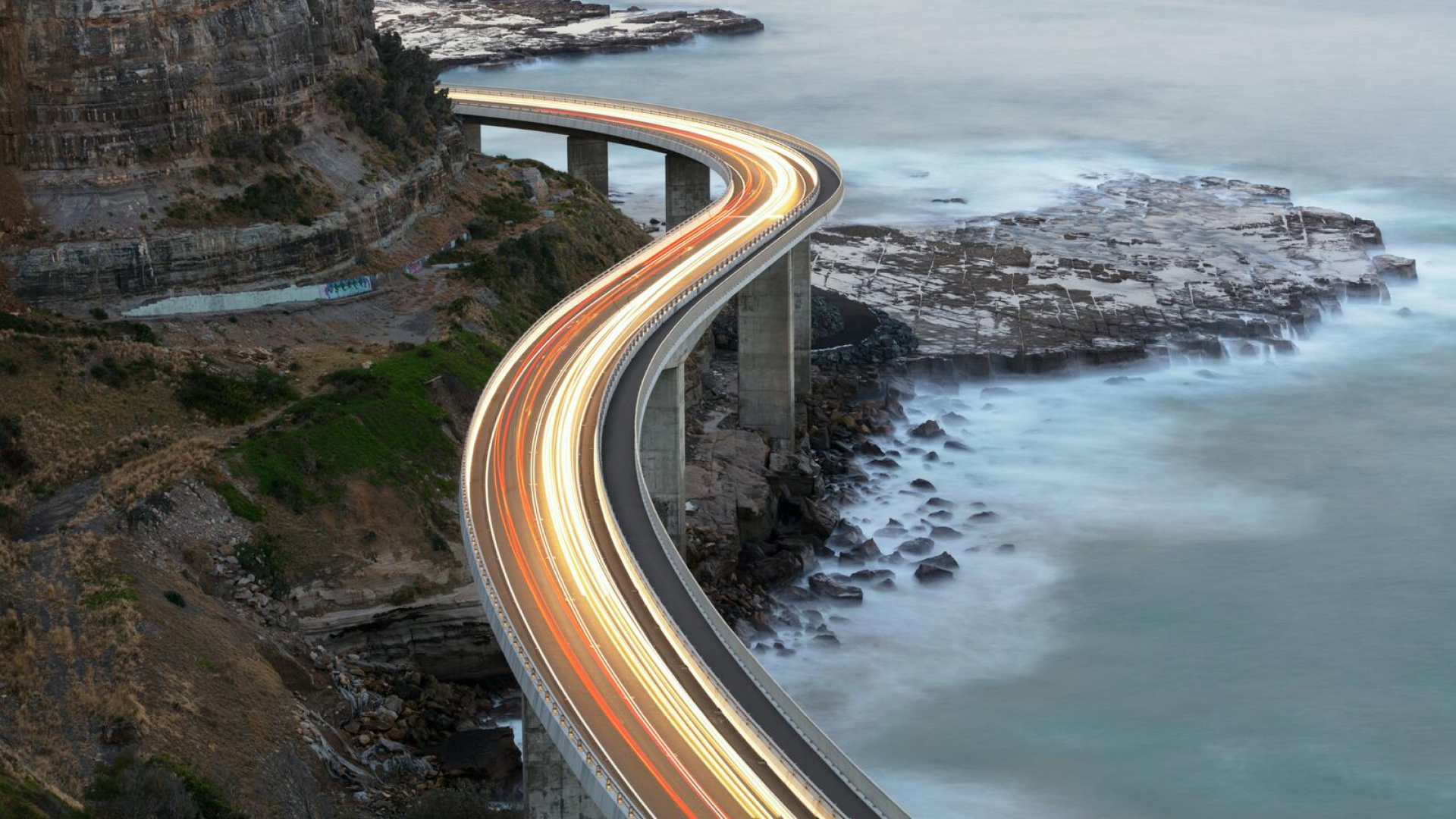 A coastal highway curves dramatically above rocky shores, with light trails from vehicles creating bright lines against the evening backdrop.